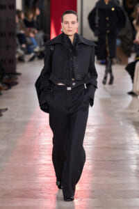 Female model walks the runway in a dark, structured utility-style jacket with wide lapels and matching wide-leg trousers; audience blurred in background.
