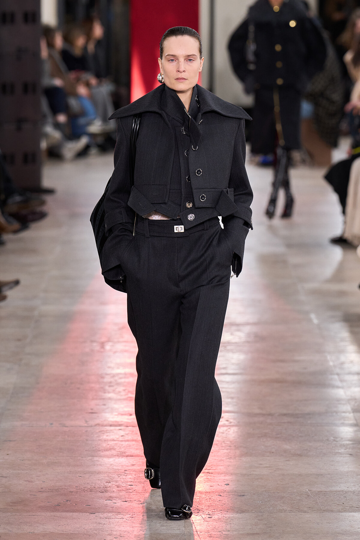 Female model walks the runway in a dark, structured utility-style jacket with wide lapels and matching wide-leg trousers; audience blurred in background.