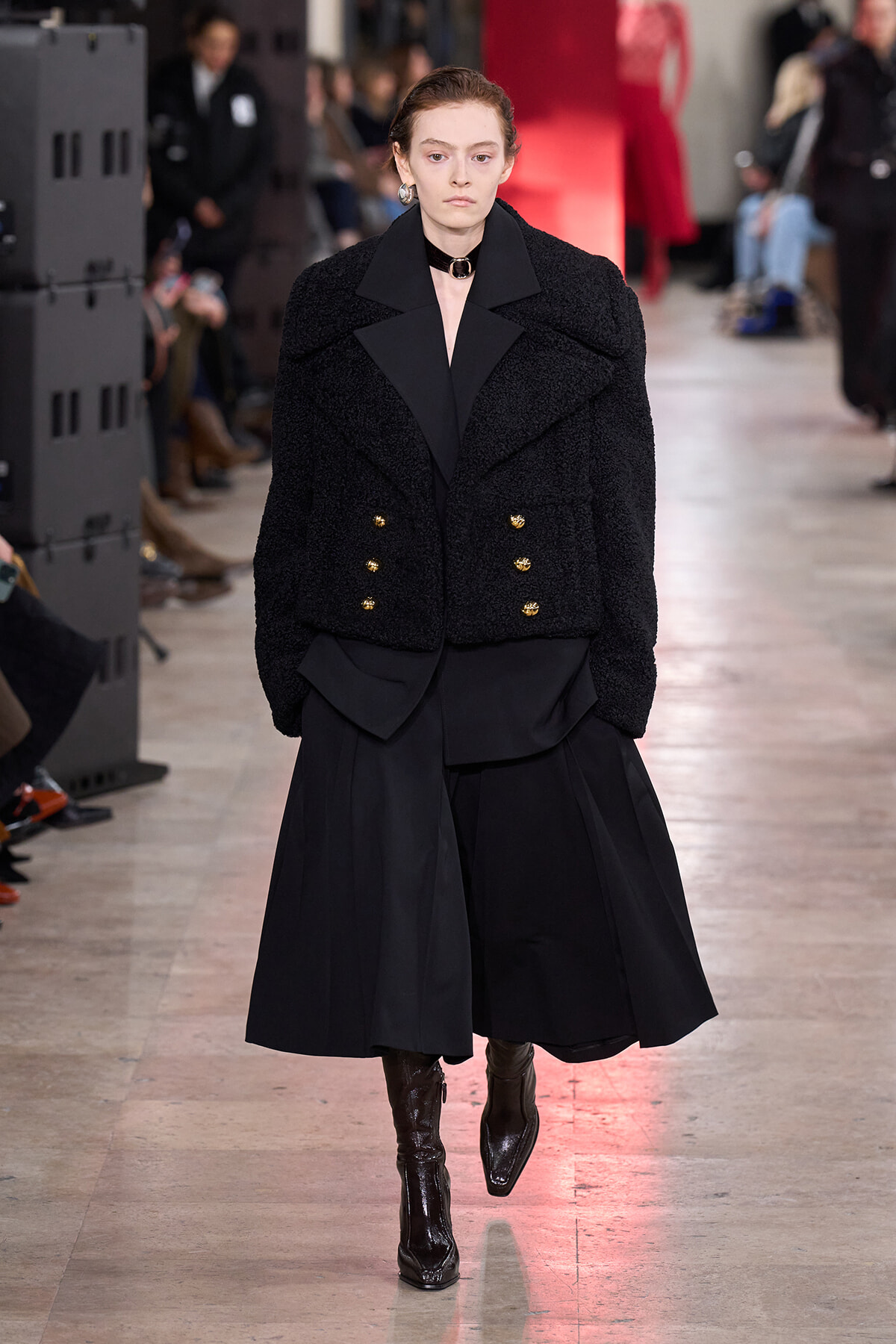 Model on a runway wearing a black boucle double-breasted jacket with gold buttons and a long black skirt, brown boots visible at the hem.