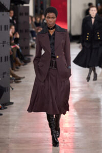 Runway model in a burgundy coordinated suit with a black shirt, statement choker, and knee-high black boots.