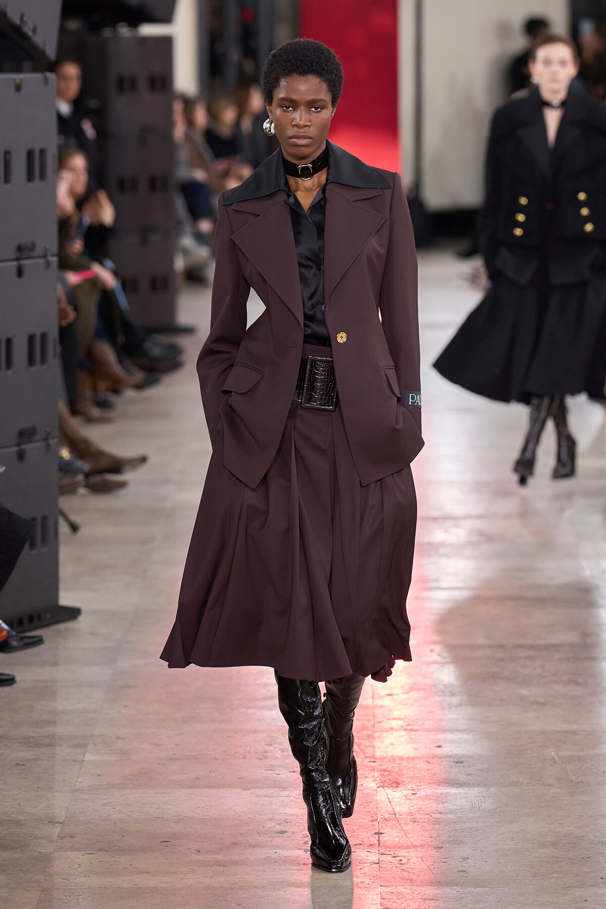 Runway model in a burgundy coordinated suit with a black shirt, statement choker, and knee-high black boots.