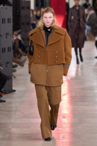 Model on a runway wearing a camel oversized double-breasted jacket with wide lapels over matching cargo pants and a small black handbag.