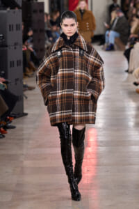 Model on a runway wearing an oversized brown plaid coat and shiny black thigh-high boots.