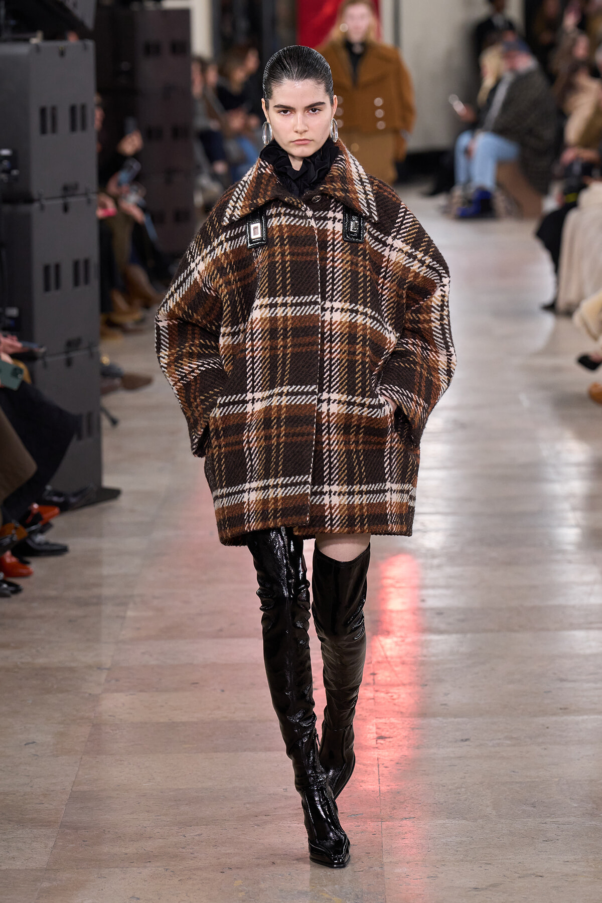 Model on a runway wearing an oversized brown plaid coat and shiny black thigh-high boots.