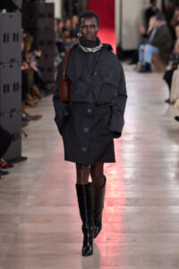 Model walking a runway in a black oversized coat with cape-like details, chunky silver necklace, brown crossbody bag, and knee-high black boots; blurred audience in background.