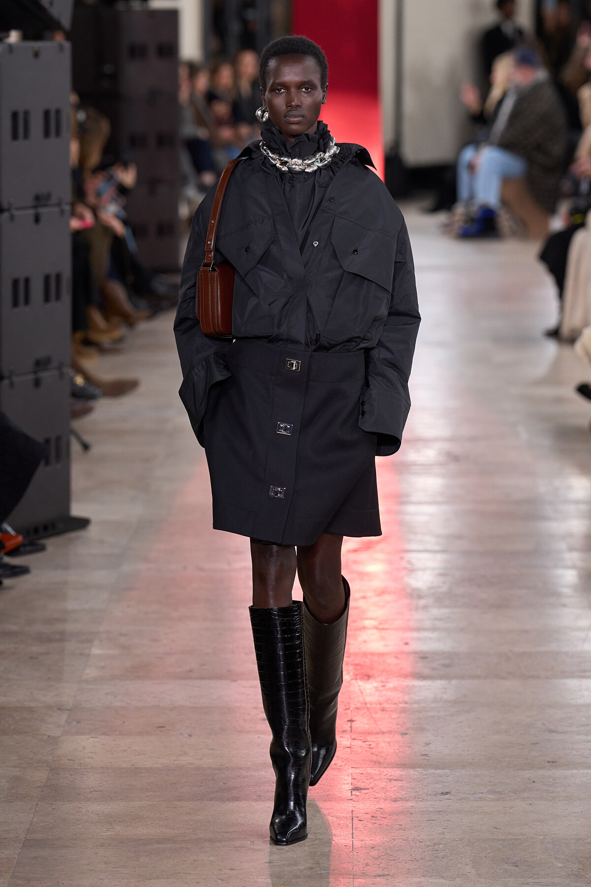 Model walking a runway in a black oversized coat with cape-like details, chunky silver necklace, brown crossbody bag, and knee-high black boots; blurred audience in background.