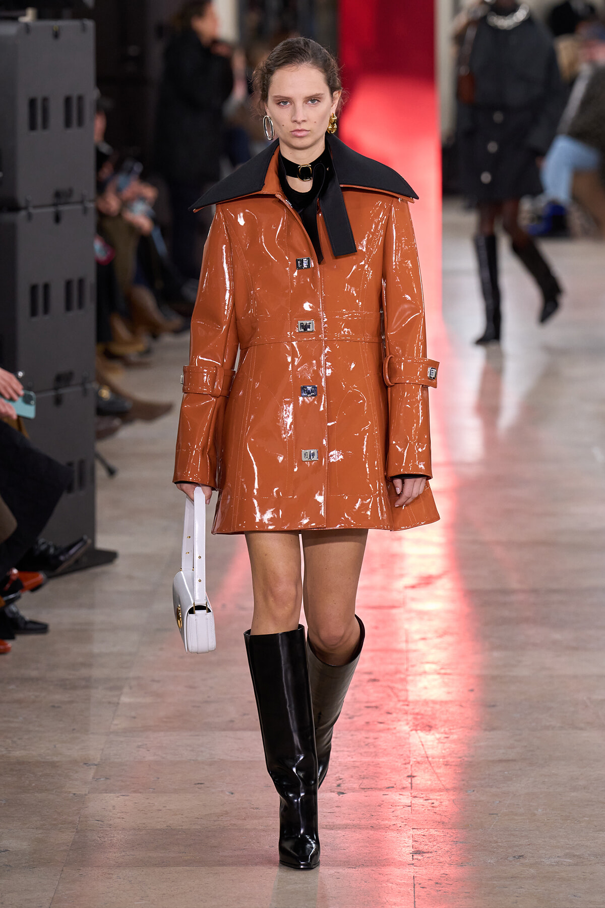 Model wearing a glossy tan-brown patent trench coat with oversized black collar, black knee-high boots, and a white handbag walking a fashion runway.