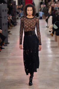 Model walks the runway in a sheer black lace long-sleeve top paired with a black lace midi skirt and a wide glossy belt, finished with tall black boots.