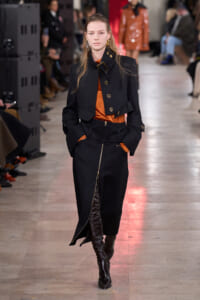 Model walking the runway in a black cropped jacket with gold buttons over an orange top, matching high-waisted black skirt and knee-high black boots.