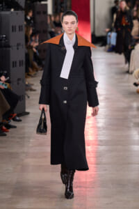 Model on a runway wearing a long black coat with orange-brown oversized shoulders, white necktie, and black heeled boots, carrying a small black handbag.