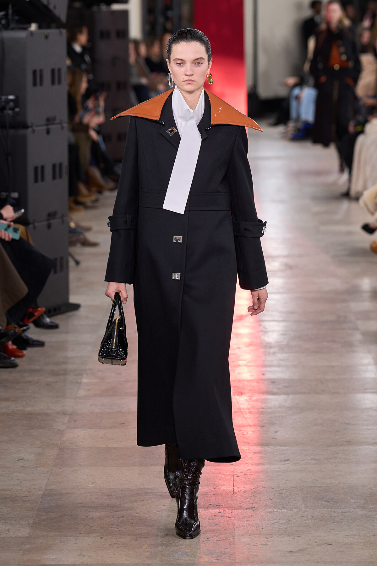 Model on a runway wearing a long black coat with orange-brown oversized shoulders, white necktie, and black heeled boots, carrying a small black handbag.