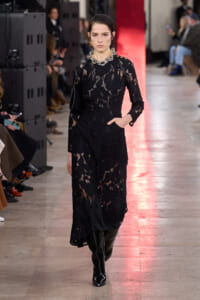 Model in a black lace maxi dress with a silver chunky necklace walks the runway, knee-high black boots and a small black bag at her side