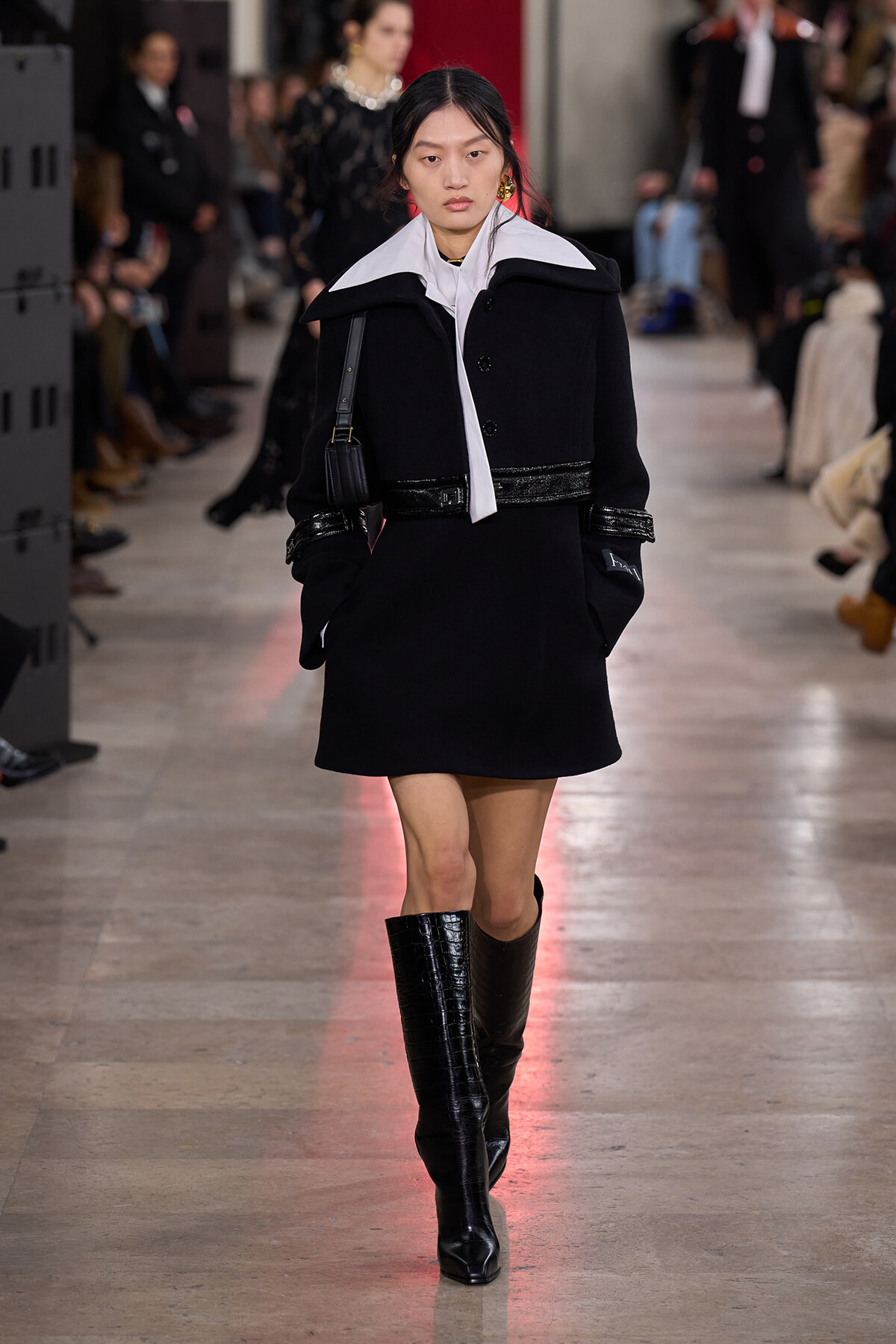 Model walking a runway in a black coat with a white oversized collar and tall black crocodile-textured boots, carrying a small shoulder bag