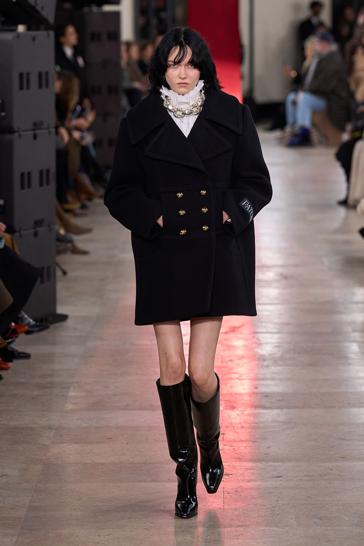 Runway model walking in a black double-breasted wool coat with gold buttons, white ruffled blouse, chunky silver necklace, and knee-high black boots.