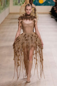 Model on a runway wearing a nude mesh gown with brown leaf appliqués and long fringe at the hem.