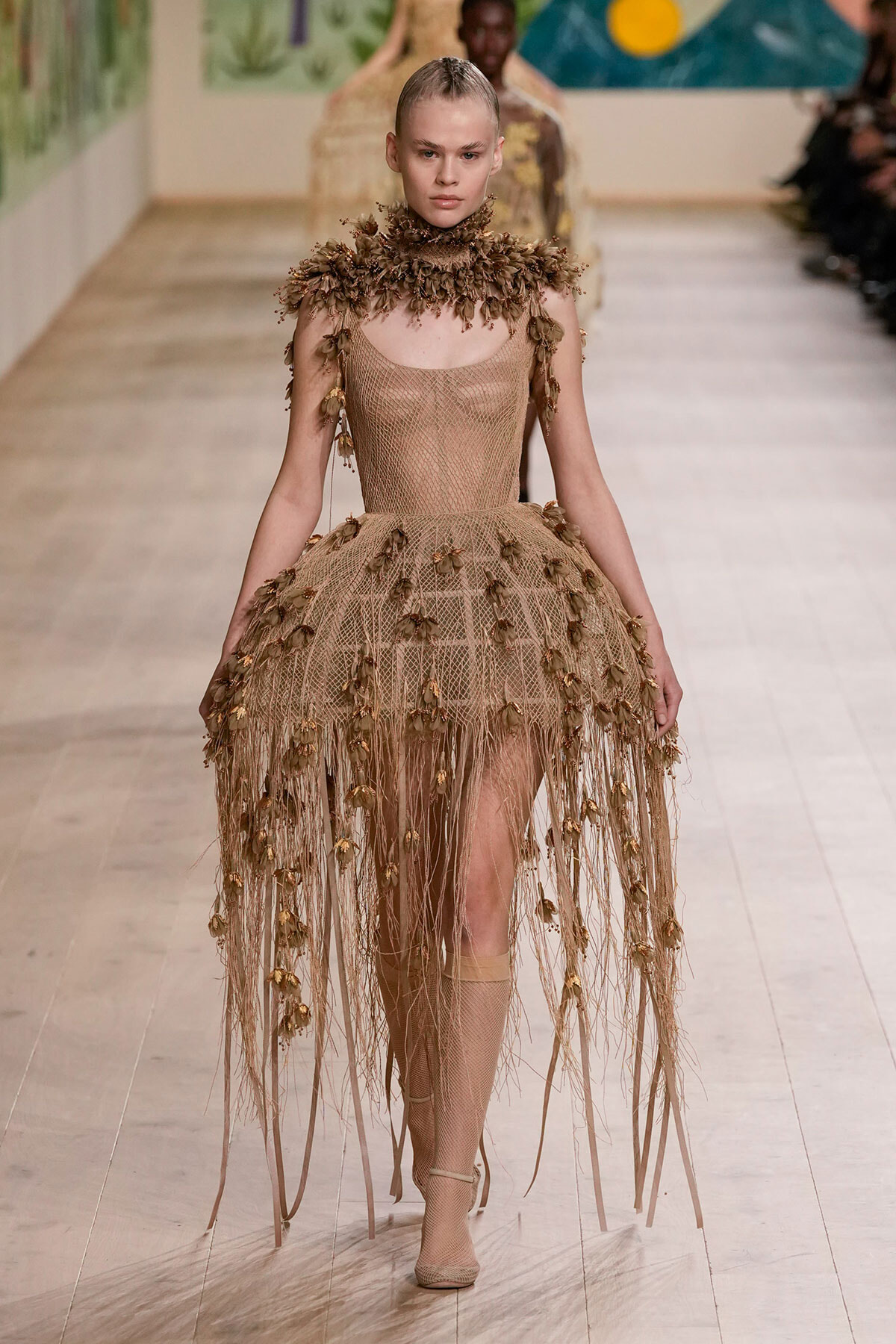 Model on a runway wearing a nude mesh gown with brown leaf appliqués and long fringe at the hem.