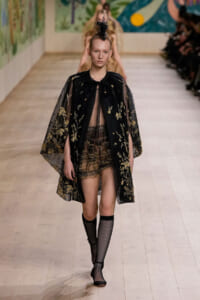 Model walks the runway in a sheer black cape with gold floral embroidery over a lace mini-dress, paired with black fishnet stockings and shoes.