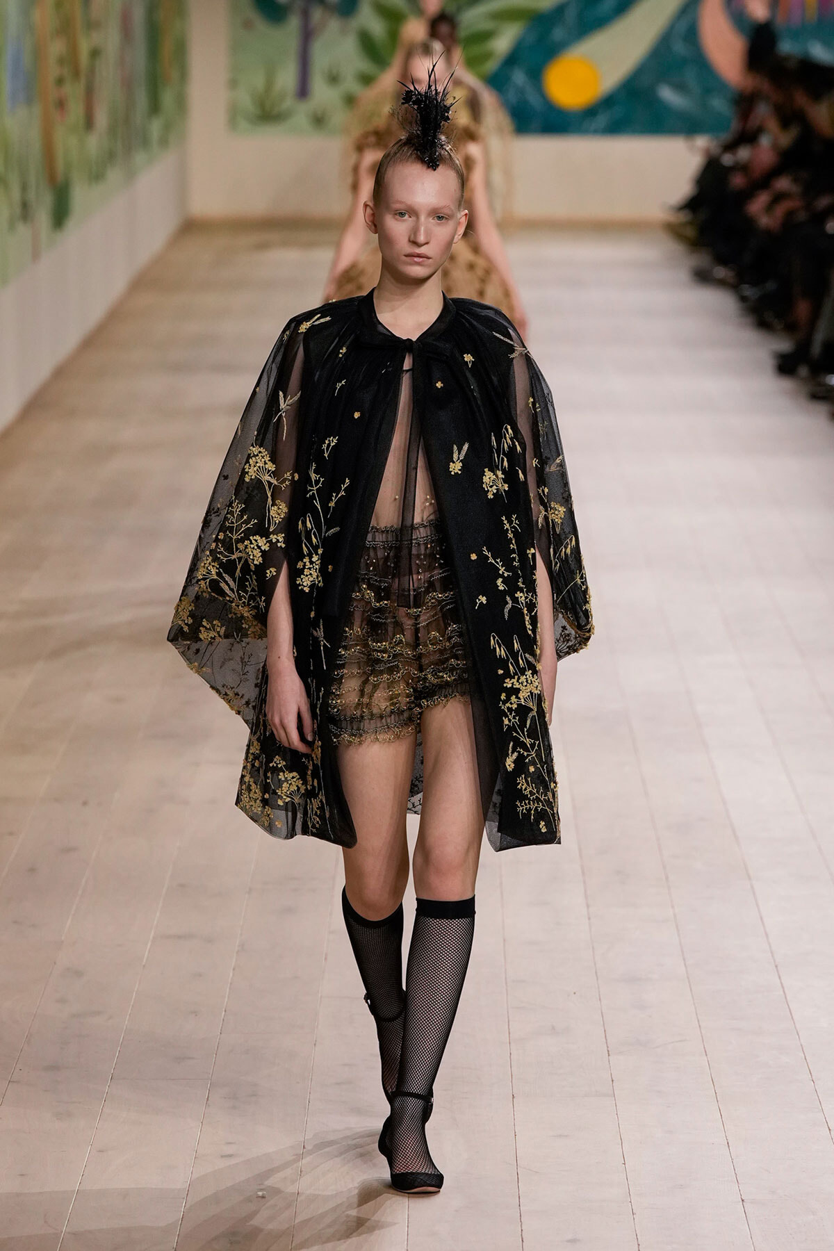 Model walks the runway in a sheer black cape with gold floral embroidery over a lace mini-dress, paired with black fishnet stockings and shoes.