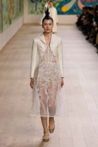 Model on a runway wearing a cream tailored blazer over a sheer lace dress with a black feathered headpiece.