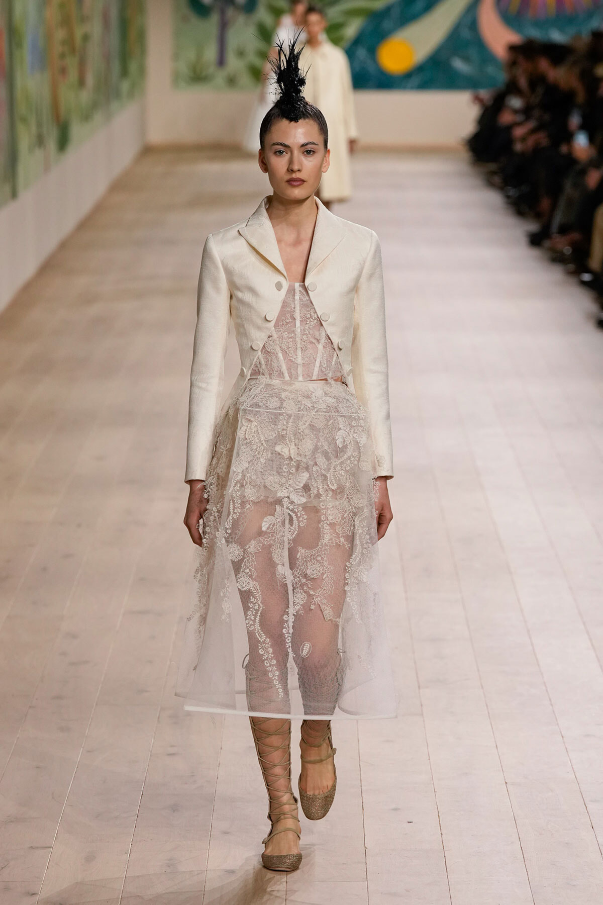 Model on a runway wearing a cream tailored blazer over a sheer lace dress with a black feathered headpiece.