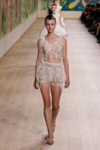 Model walks the runway in a sheer lace mini dress with floral appliqués, beige lace‑up sandals, and a black feather headpiece.