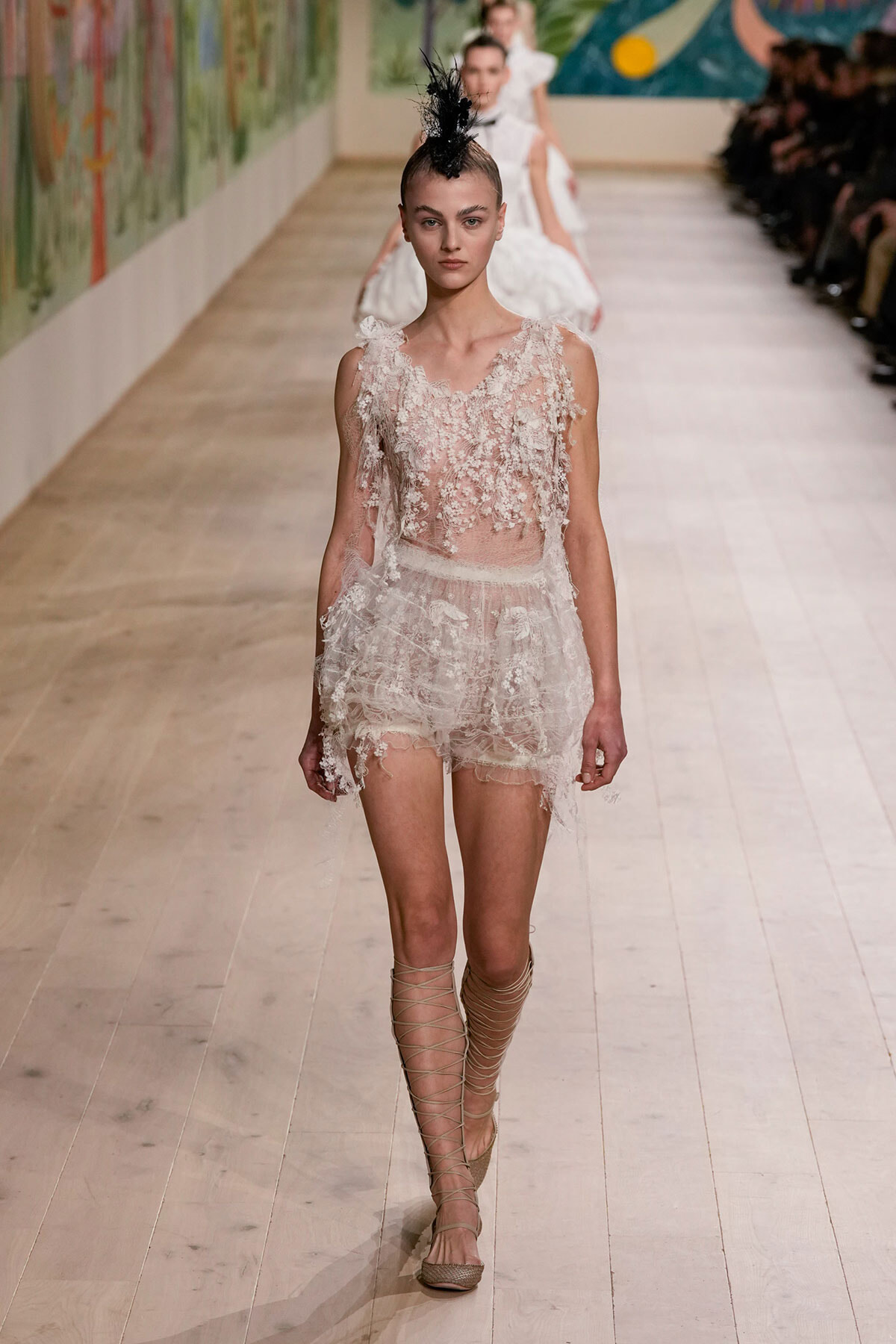 Model walks the runway in a sheer lace mini dress with floral appliqués, beige lace‑up sandals, and a black feather headpiece.