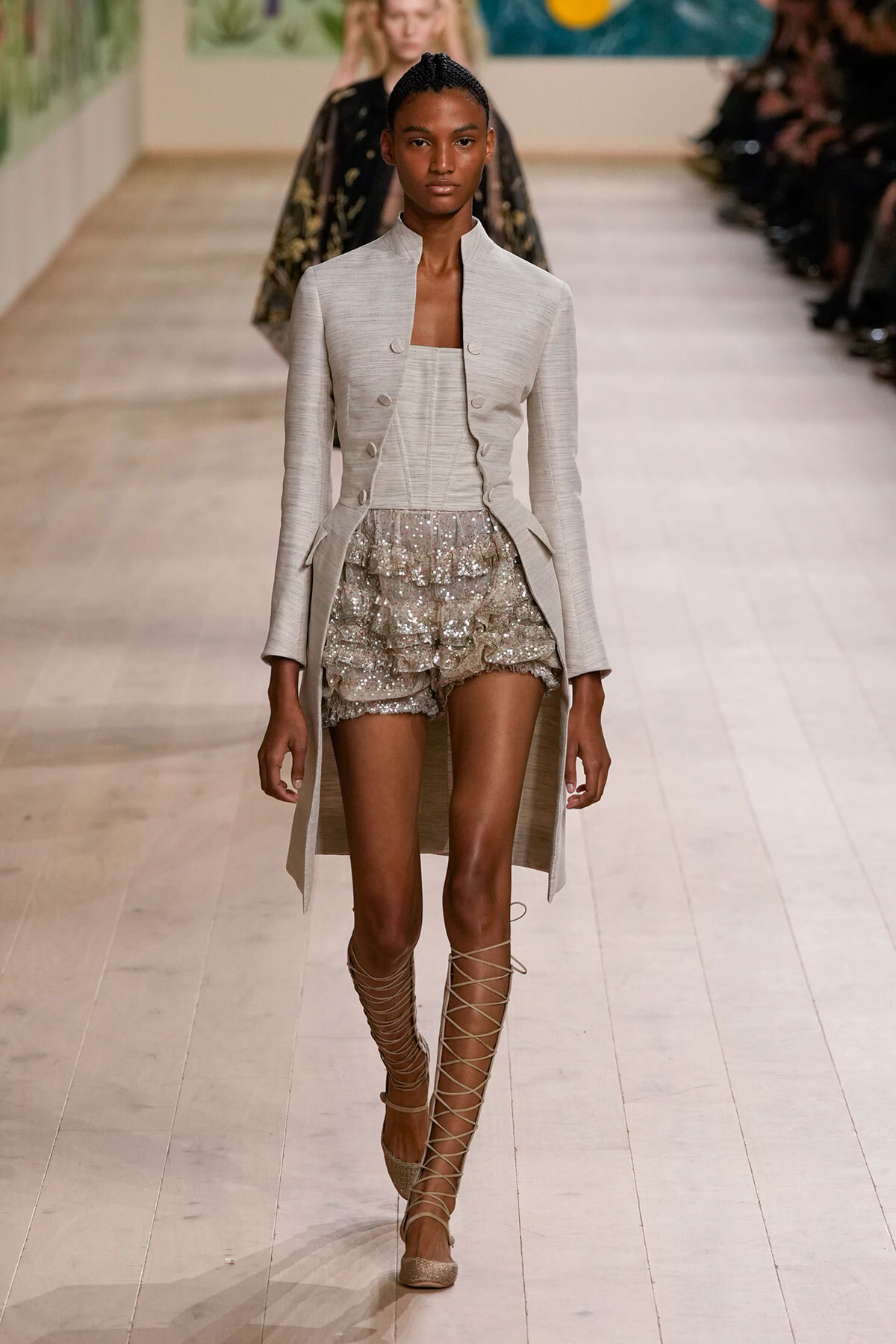 Model on a runway wearing a light gray tailored coat over a matching top and sparkling silver sequin shorts with lace-up gladiator sandals.