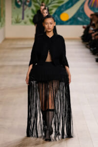 Model on a runway wearing a black textured outfit with a long fringe skirt and sheer mesh panel, hair styled in a tall spiked bun, audience in the background.