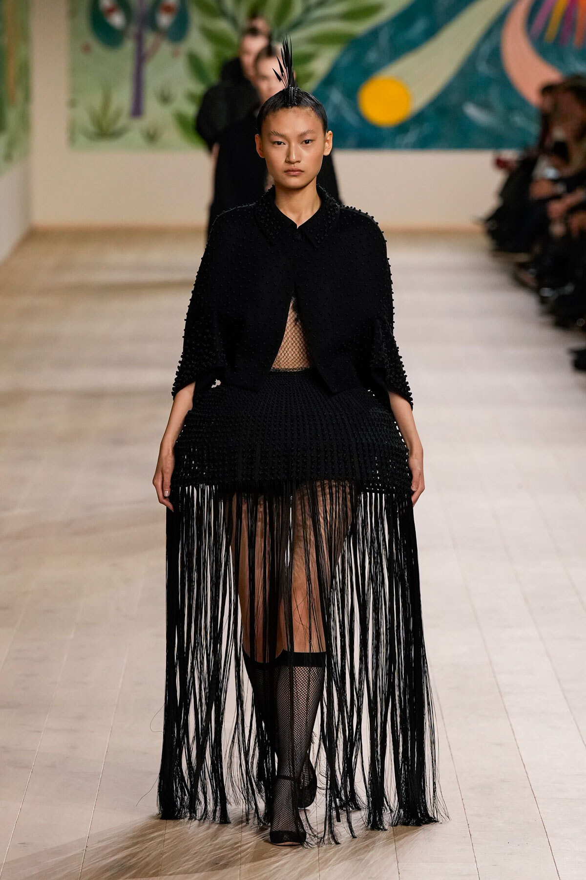 Model on a runway wearing a black textured outfit with a long fringe skirt and sheer mesh panel, hair styled in a tall spiked bun, audience in the background.