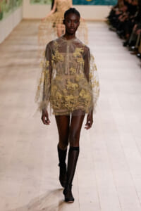 Model walking the runway in a sheer beige gown with gold floral embroidery and black fishnet socks.