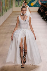 Runway model in a white lace sleeveless gown with a high-low hem, black lace-up sandals, and a black choker with a neck ribbon.