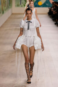 Model walks a white lace mini dress with ruffled peplum and long ribbon streamers on a fashion runway, black ribbon bow at neck, lace-up sandals, bold hair accessory