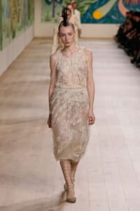 Model on a runway wearing a sheer beige lace dress with floral embroidery, lace-up sandals, and a tall black feather headpiece.