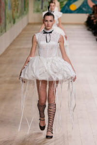 Model on a runway wearing a white lace dress with a voluminous, fringe-edged hoop skirt, a black bow choker, and black lace-up ankle-strap shoes with a feather headpiece.