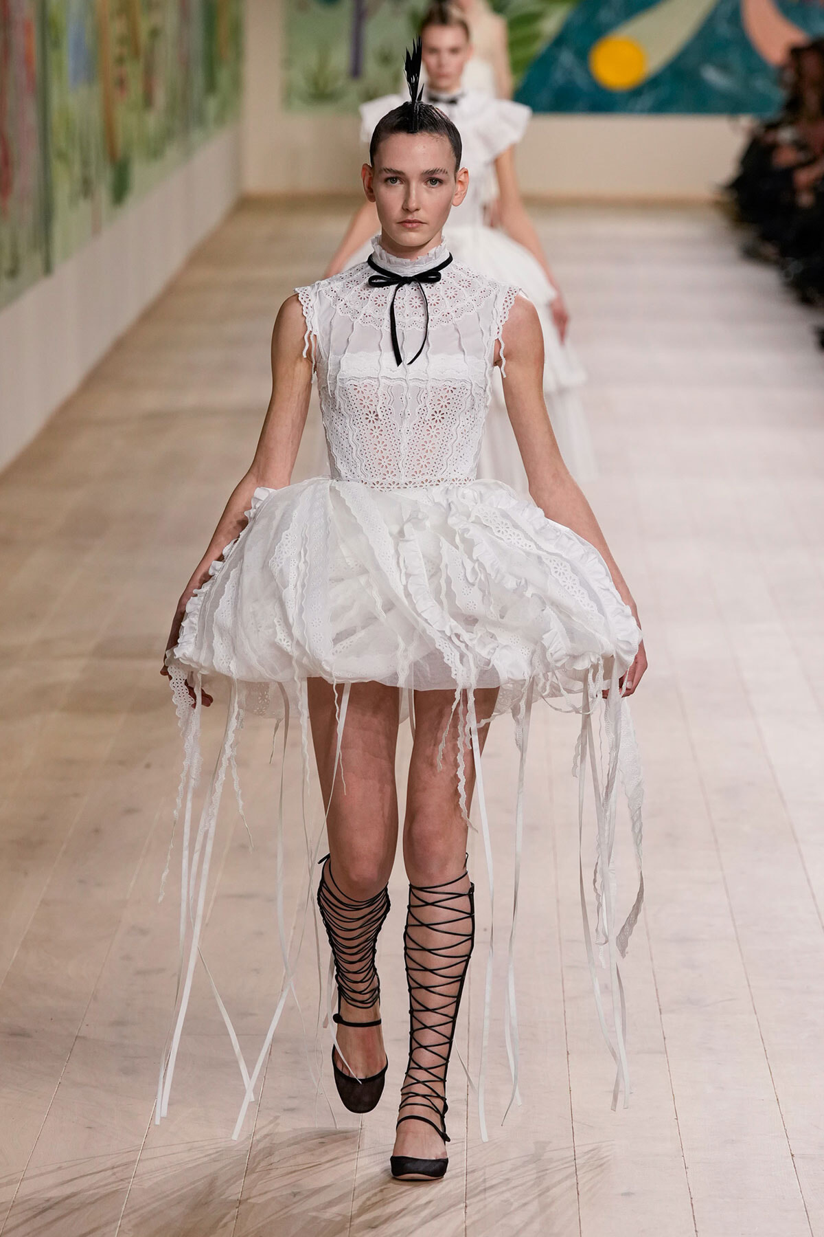 Model on a runway wearing a white lace dress with a voluminous, fringe-edged hoop skirt, a black bow choker, and black lace-up ankle-strap shoes with a feather headpiece.