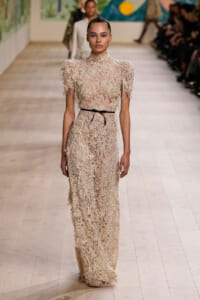 Model walks the runway in a pale cream lace gown with a high neck, short puff sleeves, and a thin black belt around the waist