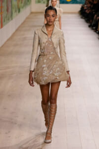 Model walking a runway in a beige lace dress with a matching cropped jacket and lace-up gladiator sandals.