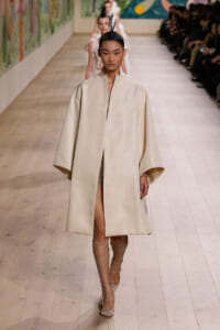 Model walks the runway in a loose cream cape coat with a minimalist design, wearing a feathered headpiece and lace-up strappy heels.