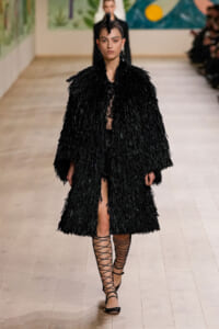 Model walks the runway in a long black feathered coat with a spiked up hairstyle, black lingerie-style outfit underneath, and crisscross lace-up sandals.
