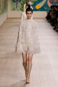Model on a runway wearing a voluminous white feathered dress and beige lace-up heels, hair in a tall feathered bun.