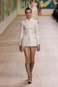 Model walks the runway in a white structured blazer-dress with peplum hips and lace shorts showing lace trim beneath. Black lace-up sandals.
