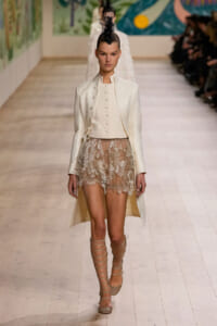 Model walks the runway in a cream tailored coat over a buttoned ivory vest and sheer lace mini skirt, with tall lace-up sandals and a black feather headpiece.