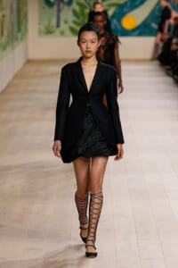 Runway model wearing a black blazer-style dress with a plunging neckline and layered skirt, paired with knee-to-thigh lace-up sandals.