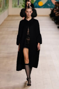 Model walks a fashion show wearing a long black textured coat with bead details over sheer lace and black lingerie, paired with fishnet stockings and heels.