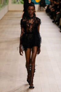 Model walking a runway in a sheer black lace mini dress with floral appliqués and matching lace-up sandals.