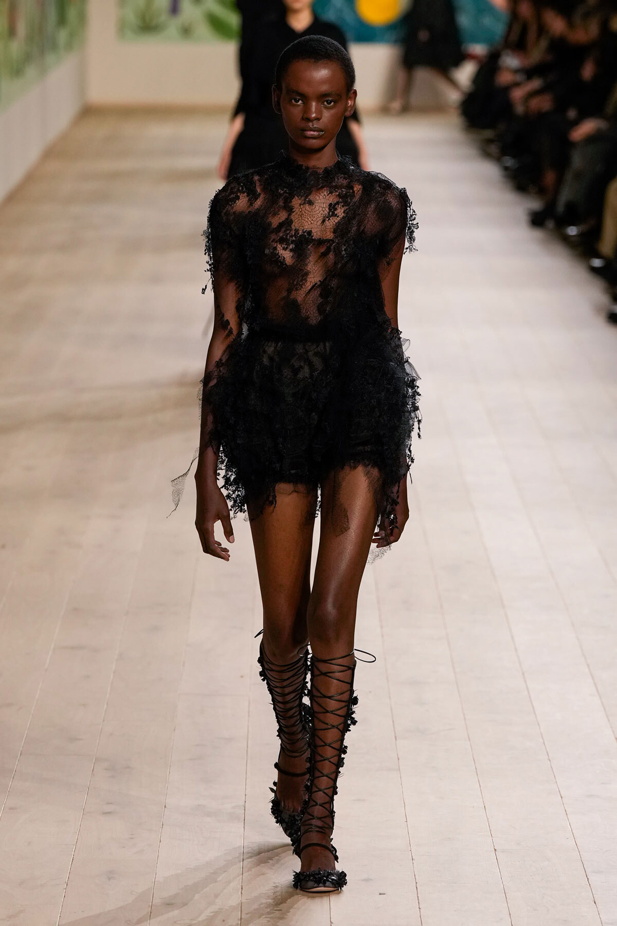 Model walking a runway in a sheer black lace mini dress with floral appliqués and matching lace-up sandals.