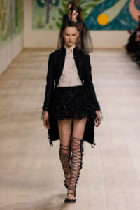Model on a runway wearing a black embroidered coat over a pale pink buttoned vest, black lace mini skirt, and tall lace-up gladiator sandals with a feather headpiece.