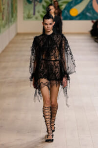 Model walks a runway in a sheer black lace outfit with dramatic puffed sleeves and lace-up sandals.