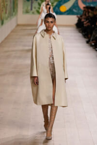 Model walks the runway in a beige oversized coat over a sheer lace dress, paired with strappy knee-high sandals.