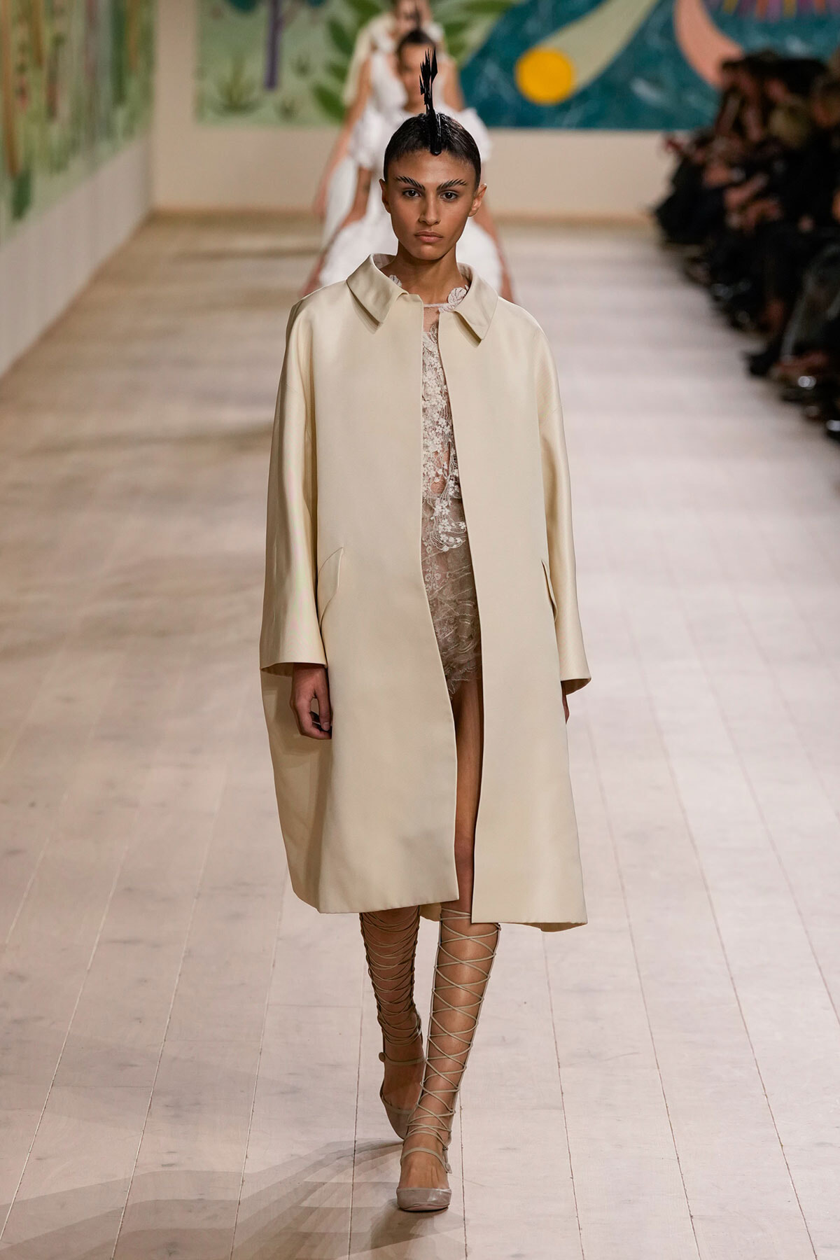 Model walks the runway in a beige oversized coat over a sheer lace dress, paired with strappy knee-high sandals.